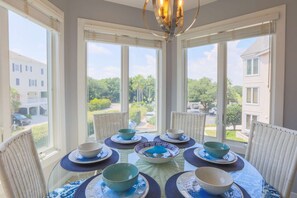 A dining area with a round table set for six, surrounded by wicker chairs, with a view of a residential neighborhood through large windows. - See, Feel & Hear the Ocean from this Light-Filled Condo w/ Community Pool & Private Screened Porch. (Isle of Palms)