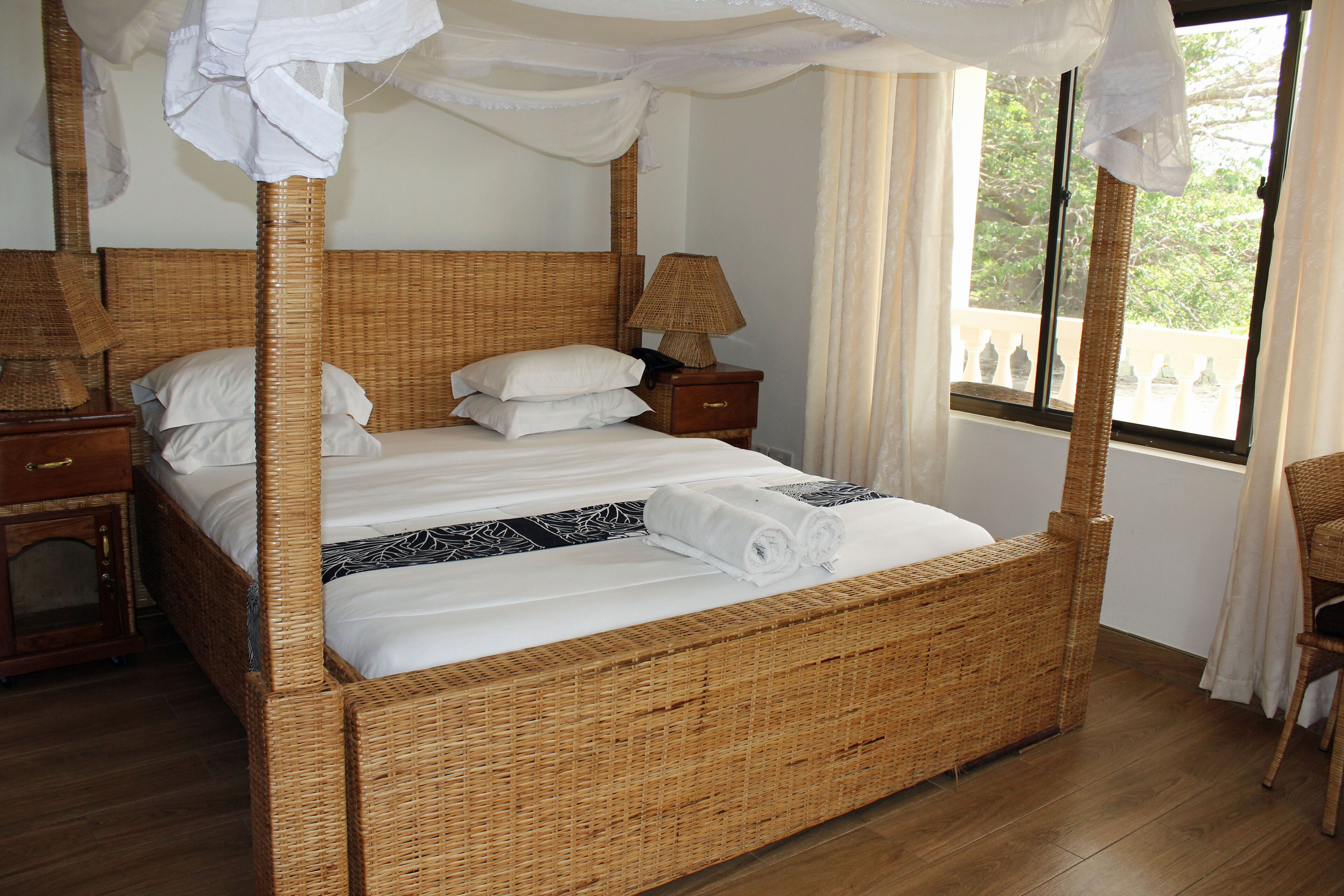 A four-poster bed with a canopy, a bedside table with a lamp, and a view of the outdoors through a window.
