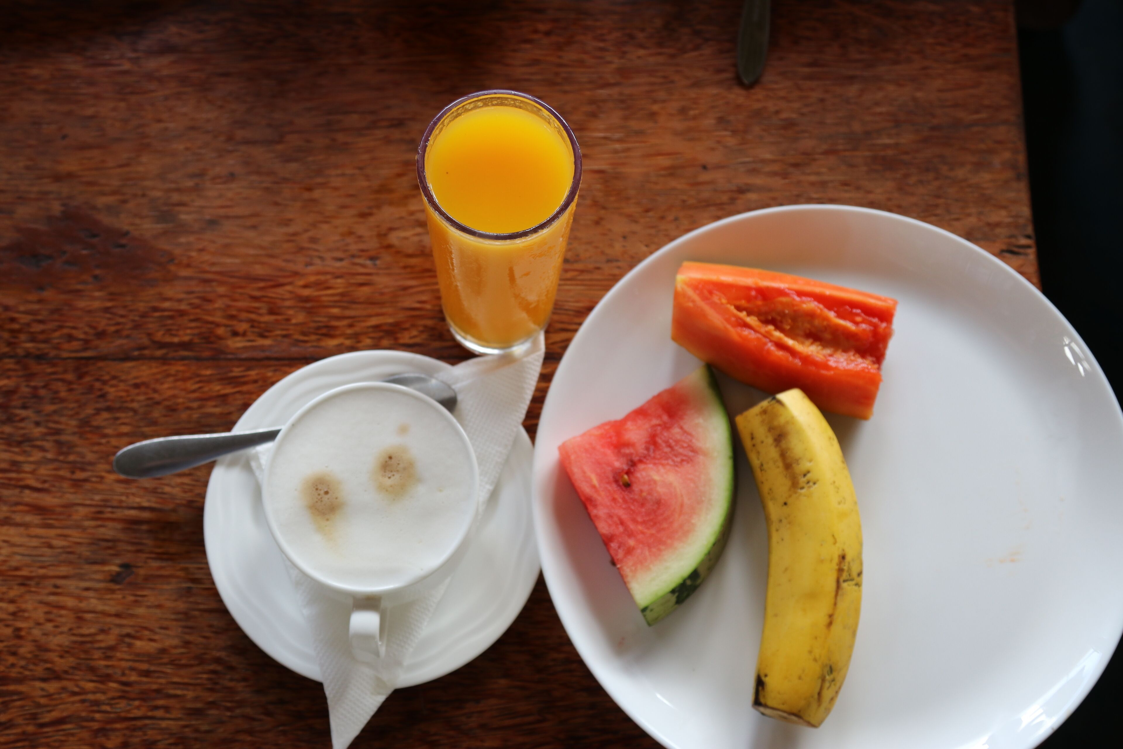 Un desayuno con una taza de café, un vaso de jugo de naranja, un plato con una rebanada de sandía y un plátano.