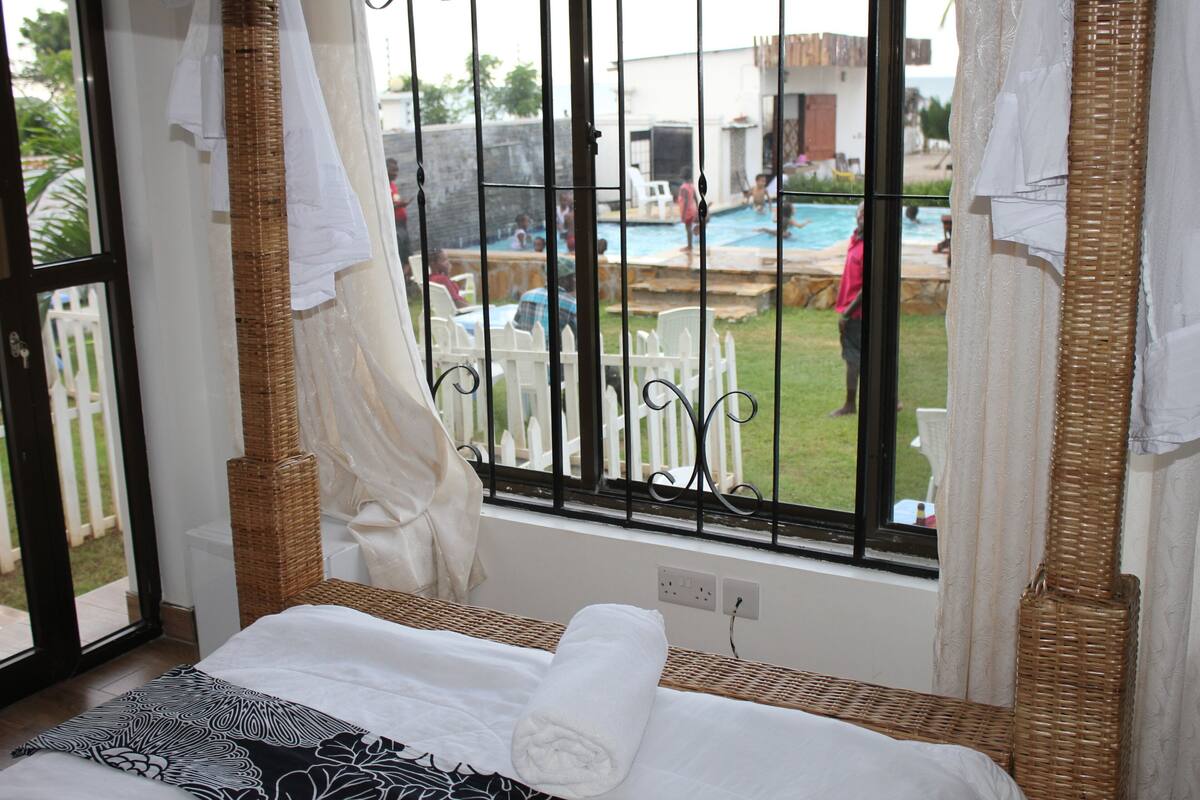 Una stanza con un letto, una finestra che si affaccia su una piscina e persone all'esterno, e una sedia in vimini.