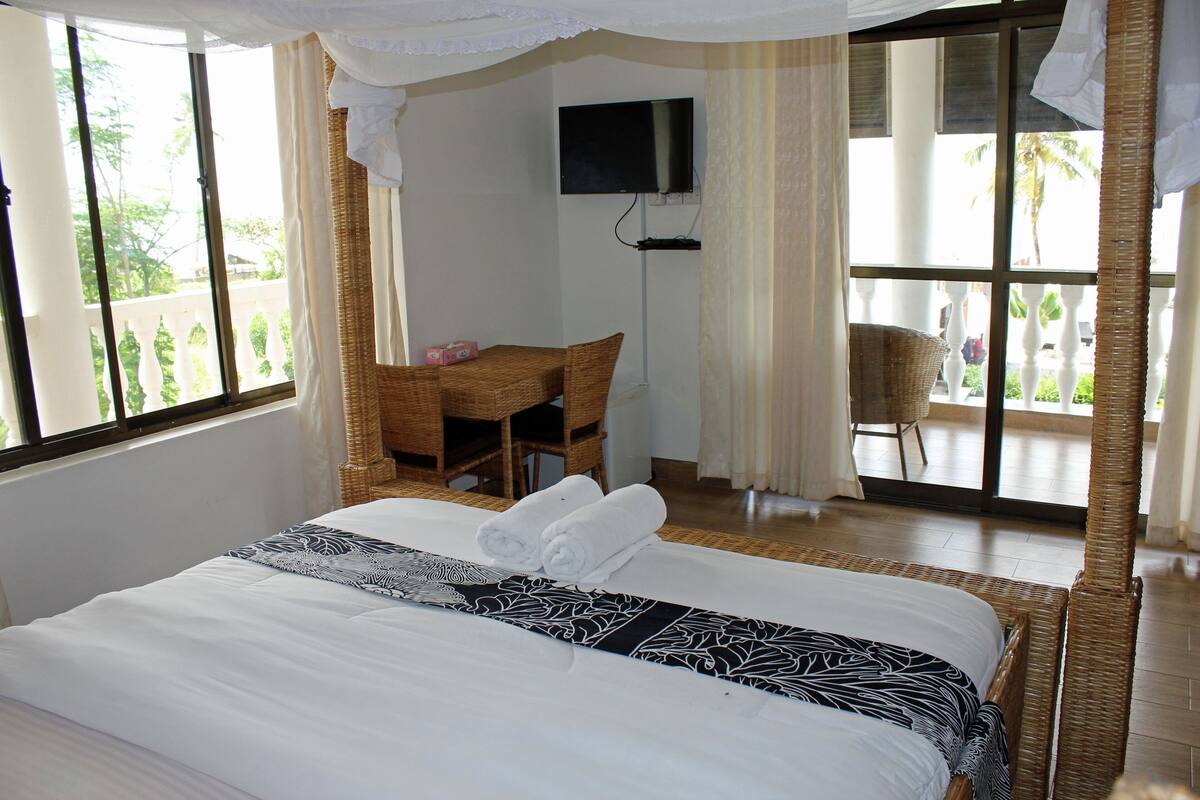 Una camera d'albergo con un letto grande, una televisione fissata al muro, una poltrona in vimini e un balcone con vista su palme.