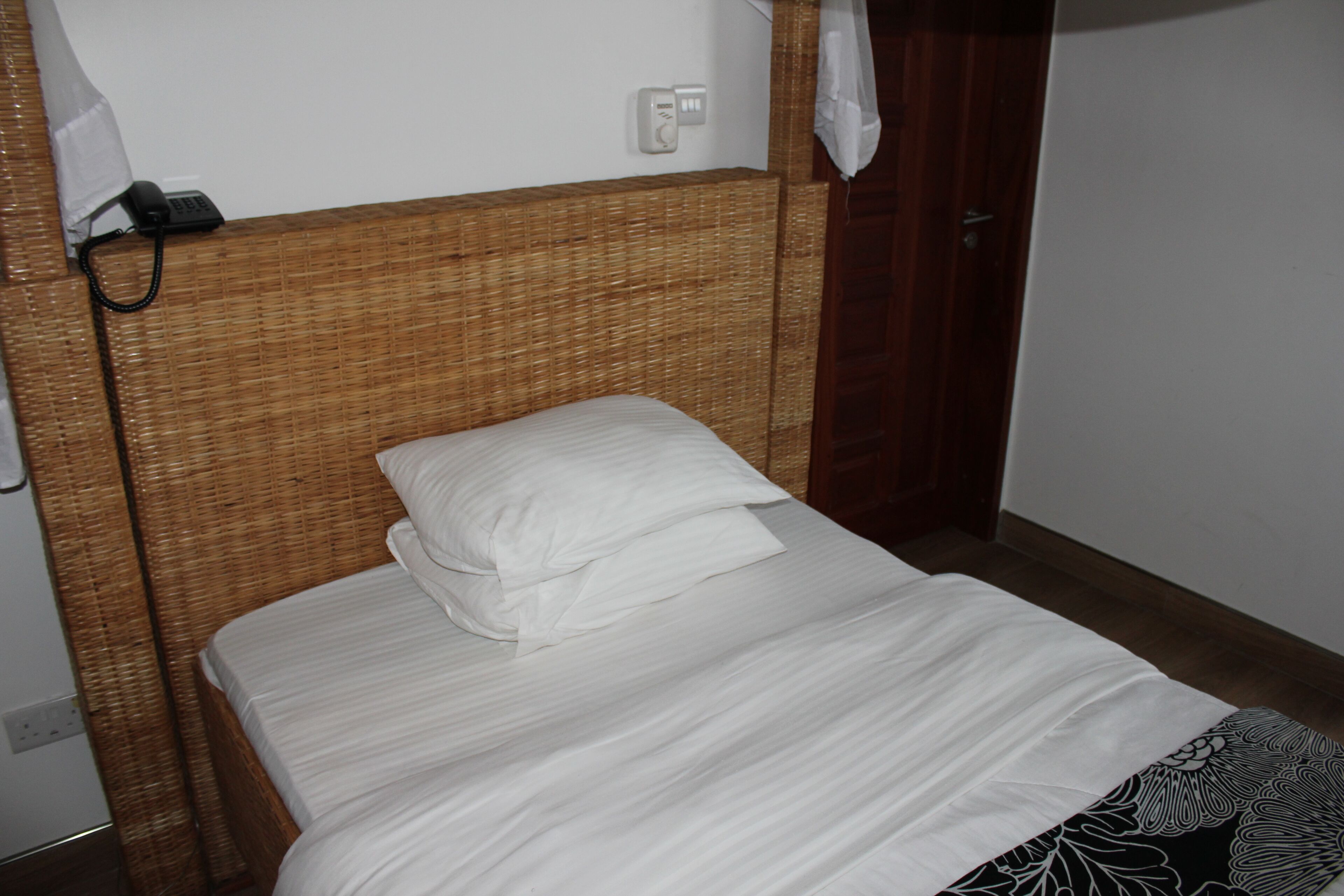 Una cama individual con ropa blanca en una habitación con cabecera de mimbre y puerta de madera.