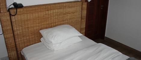 Una cama individual con ropa blanca en una habitación con cabecera de mimbre y puerta de madera.