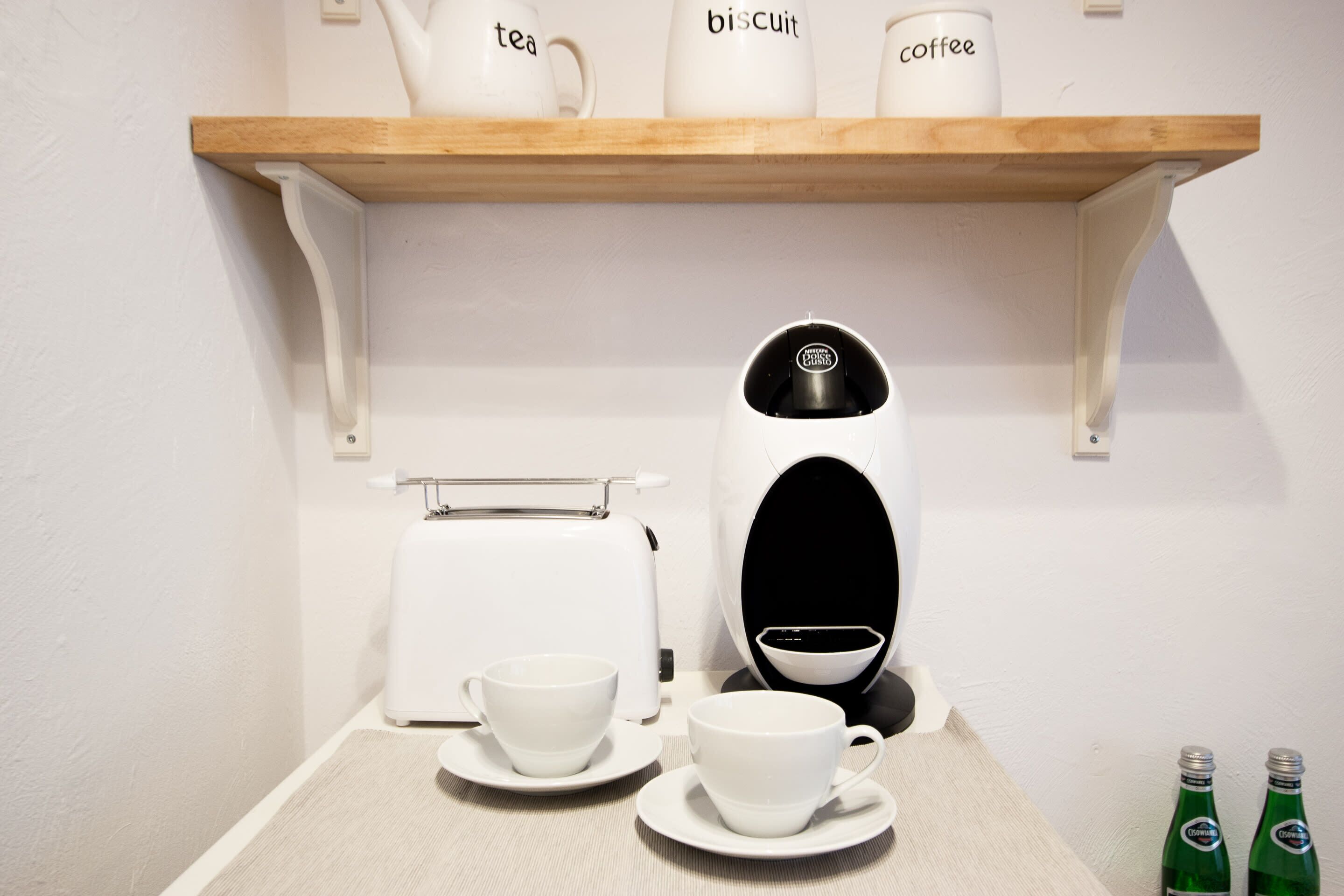 Una kitchenette con cafetera, dos tazas, un tostador y una repisa con tazas etiquetadas.