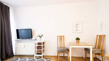 Una habitación pequeña y ordenada con televisión, un armario blanco con cestas de mimbre, dos sillas de madera, una mesita y una alfombra gris estampada.