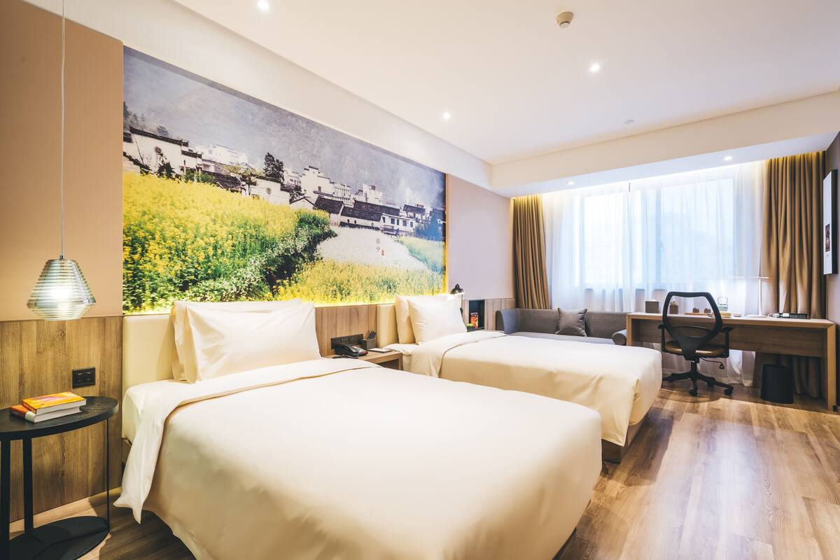 A hotel room with two beds, a desk, a chair, a large painting on the wall, and a window with curtains.