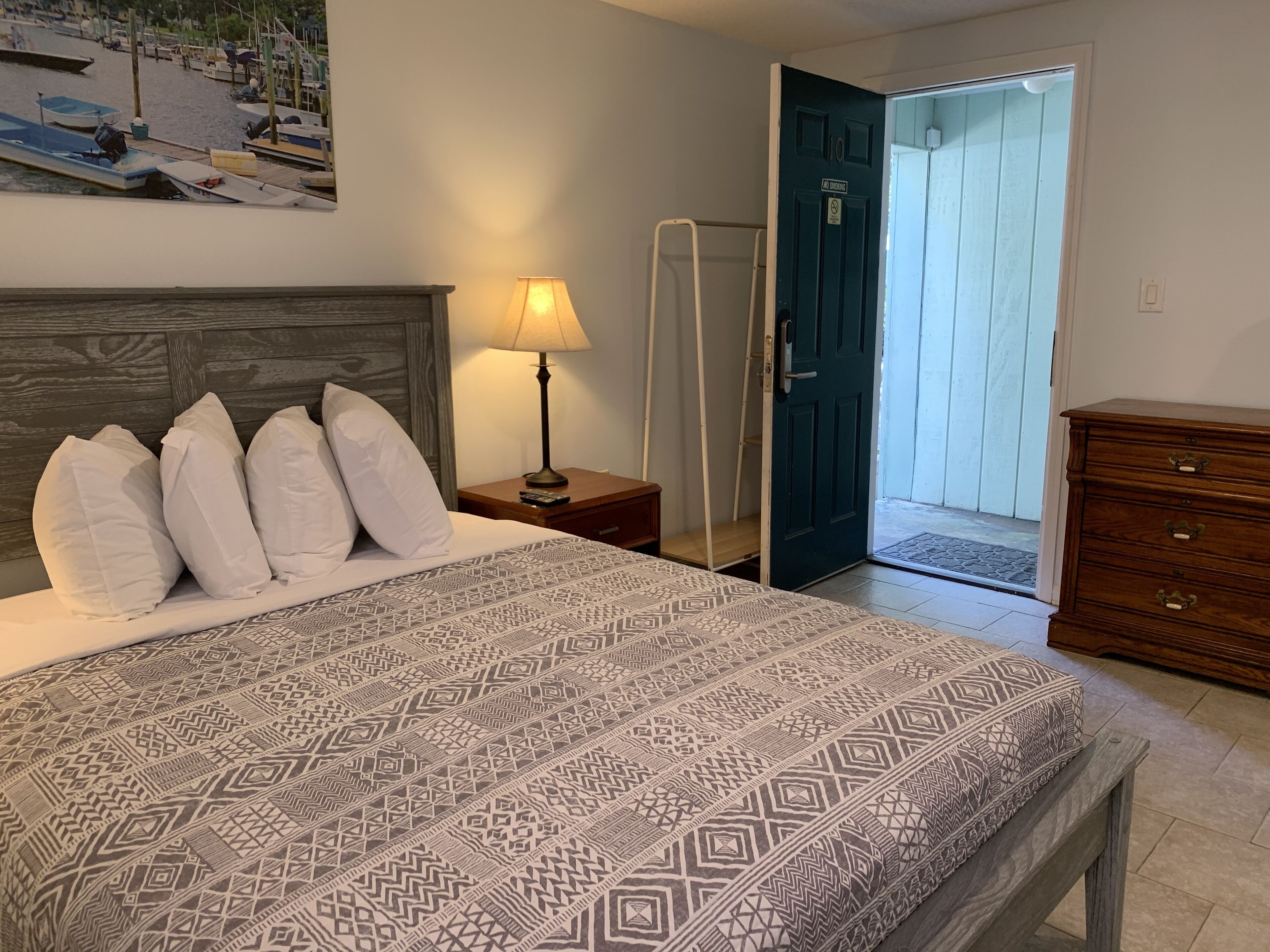 A bedroom with a patterned bedspread, a nightstand with a lamp, a wooden dresser, and a door leading to a balcony.