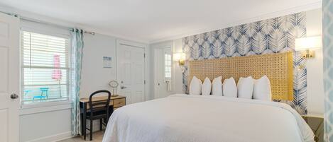Quarto com cama grande, cabeceira de madeira, mesa de cabeceira, cadeira, escrivaninha e janela com persianas.