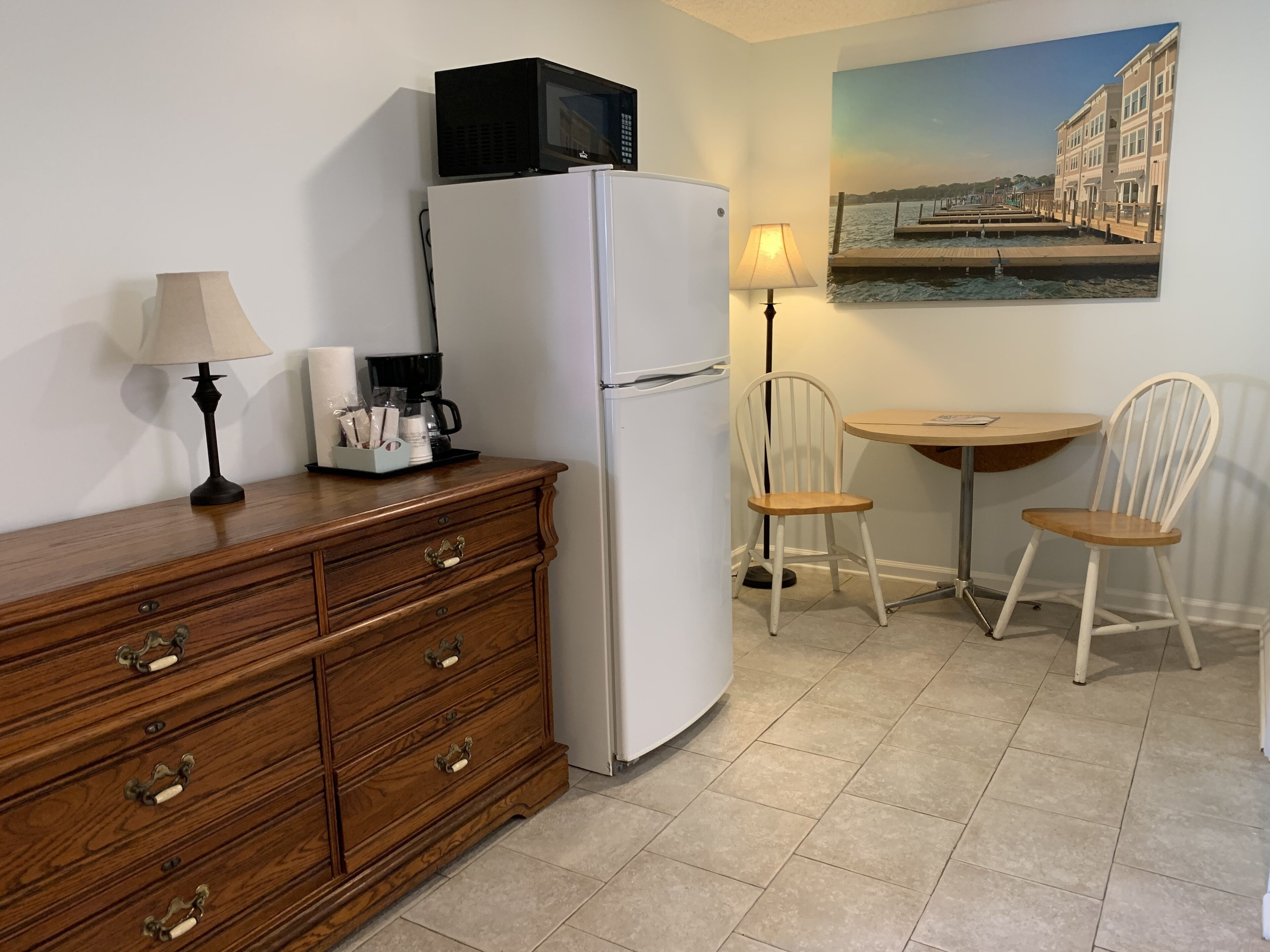A compact kitchen with a refrigerator, a wooden dresser, a coffee maker, and a table with two chairs.