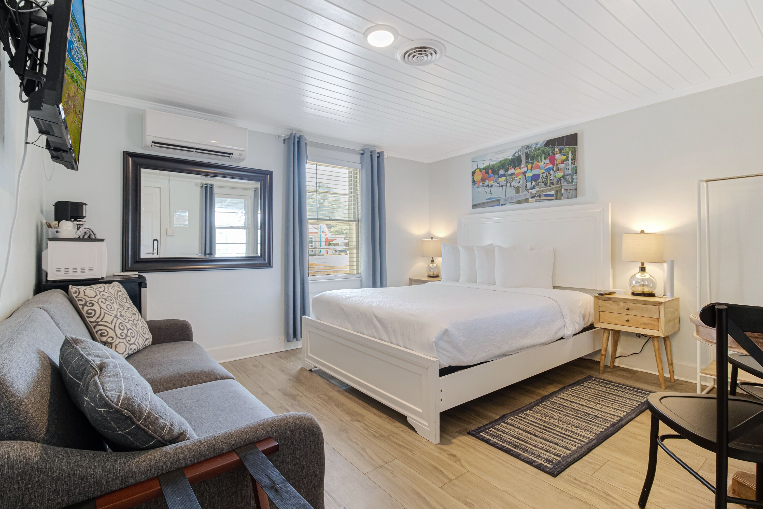 Ein Schlafzimmer mit einem Bett, einem Schlafsofa, einem Sessel, einem Schreibtisch, einer Lampe, einem Gemälde und einem Fenster mit Vorhängen.