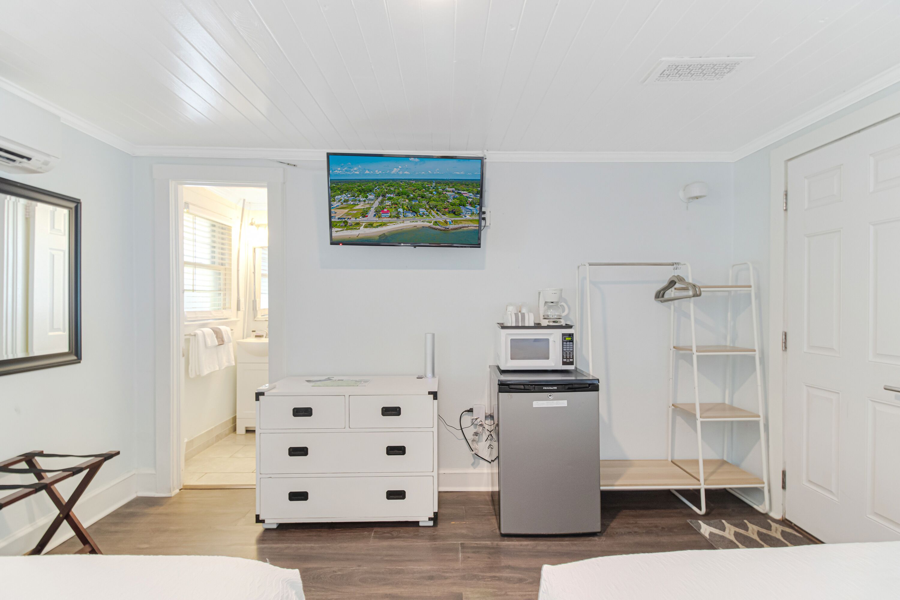A hotel room with a bed, a desk, a white dresser, a refrigerator, a microwave, a TV, and a door leading to a bathroom.