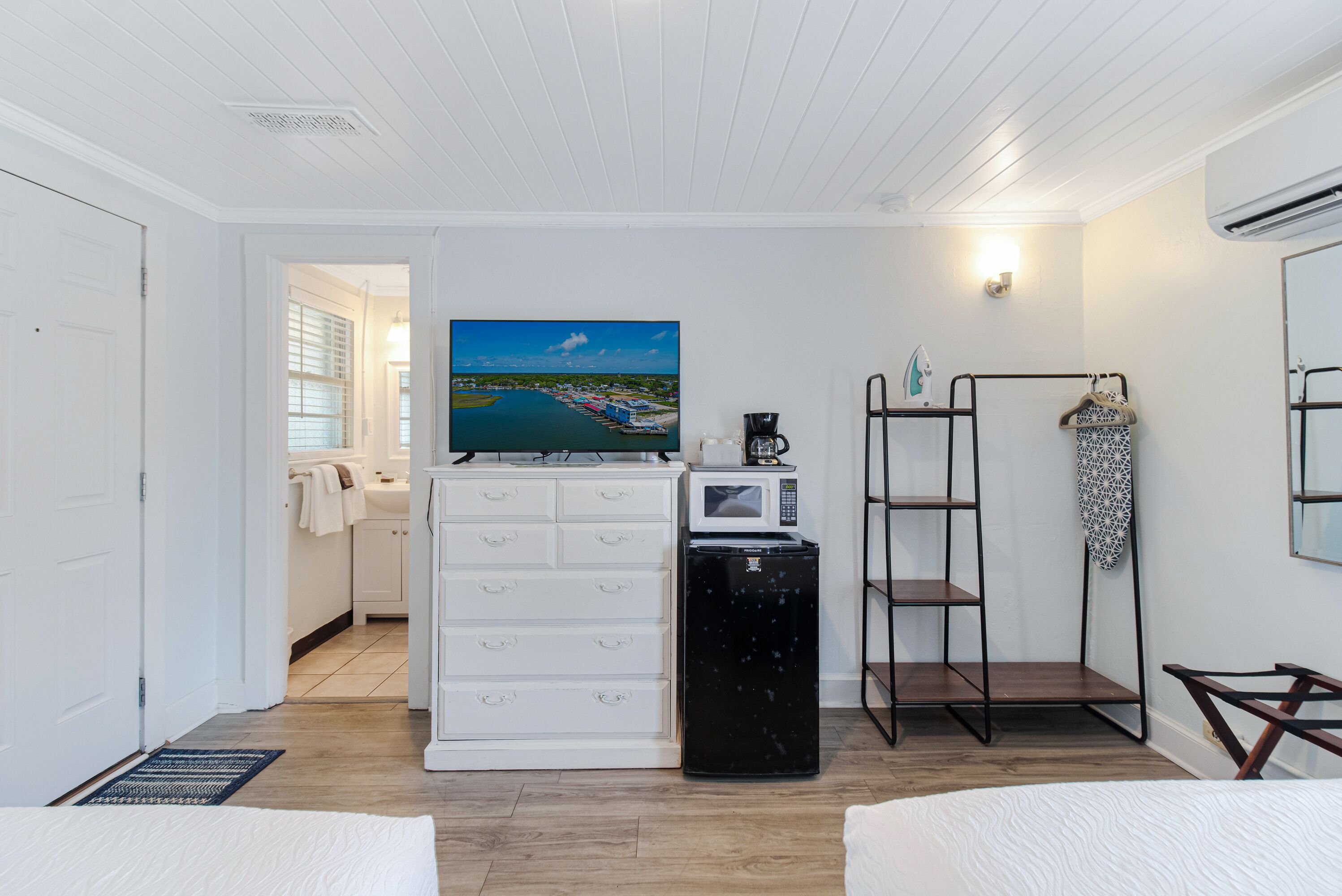 A hotel room with a bed, a TV mounted on a dresser, a mini-fridge, a microwave, a shelf with decorative items, and a view of the outside.