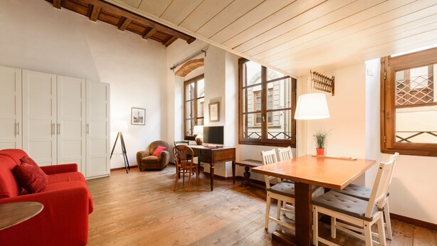 Amplia sala de estar con piso de madera, un sofá rojo, una mesa de comedor con sillas y un ventanal que proporciona luz natural.