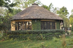 A thatched-roof house with large windows, surrounded by dense greenery. - Rhino Lodge Bardia (Thakudwara)