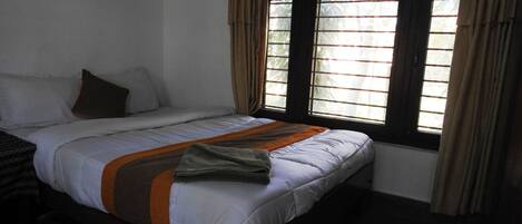 A single bed with a wooden frame, white bedding, and a brown and orange blanket. There are two windows with curtains, and a small plant is visible on the windowsill.