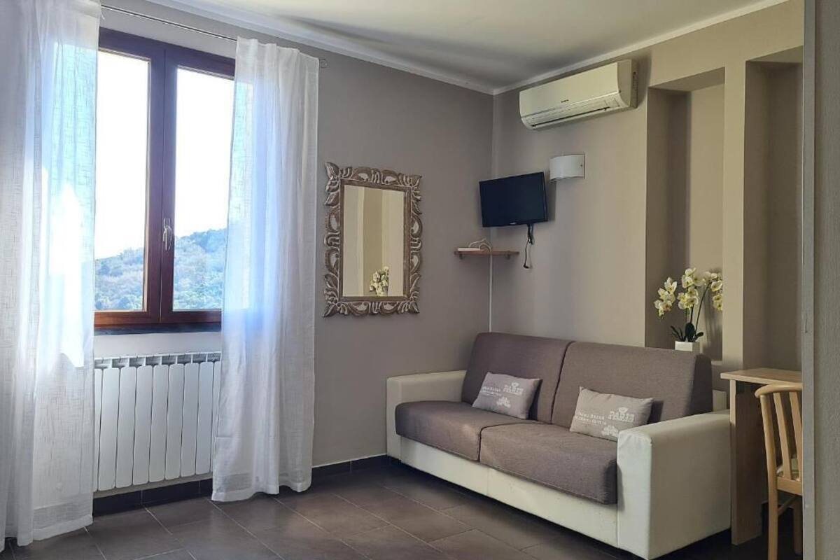 Ein Wohnzimmer mit einer Couch, einem an der Wand befestigten Fernseher, einem Fenster mit Vorhängen und einer Heizung.
