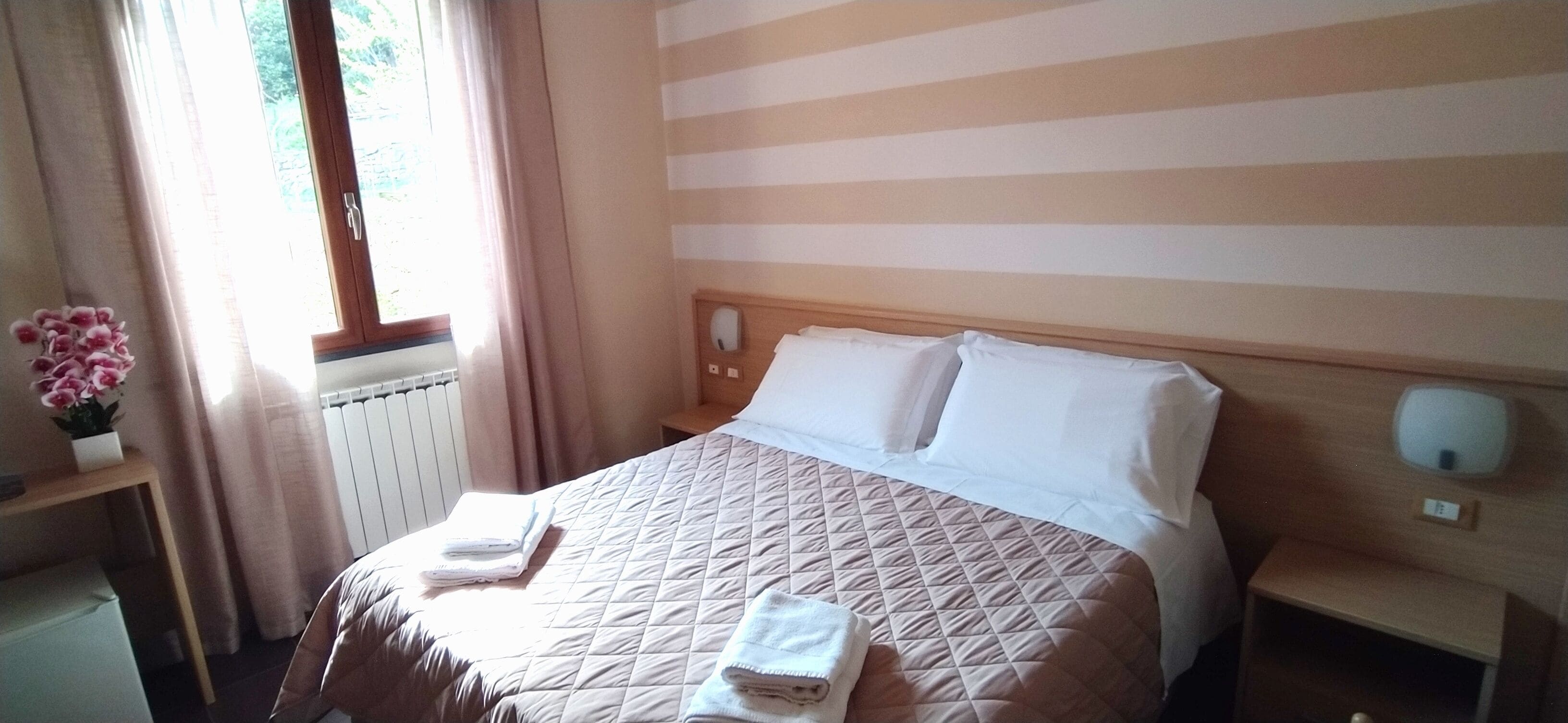 A hotel room with a bed, a bedside table, a nightstand, a window with curtains, and a small table with a vase of flowers.