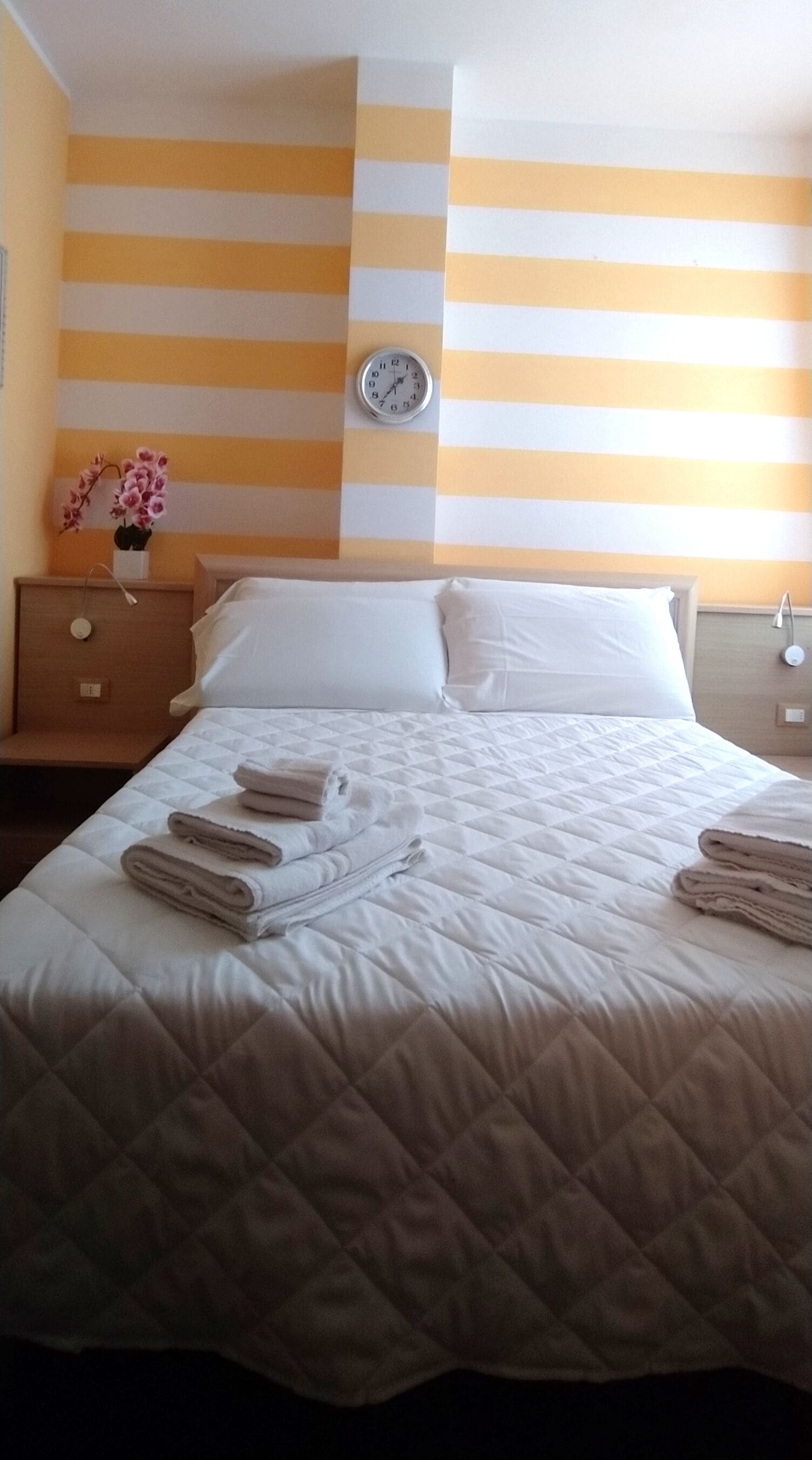 Una camera d'albergo con un letto, cuscini, una sveglia, un comodino e un vaso con fiori.