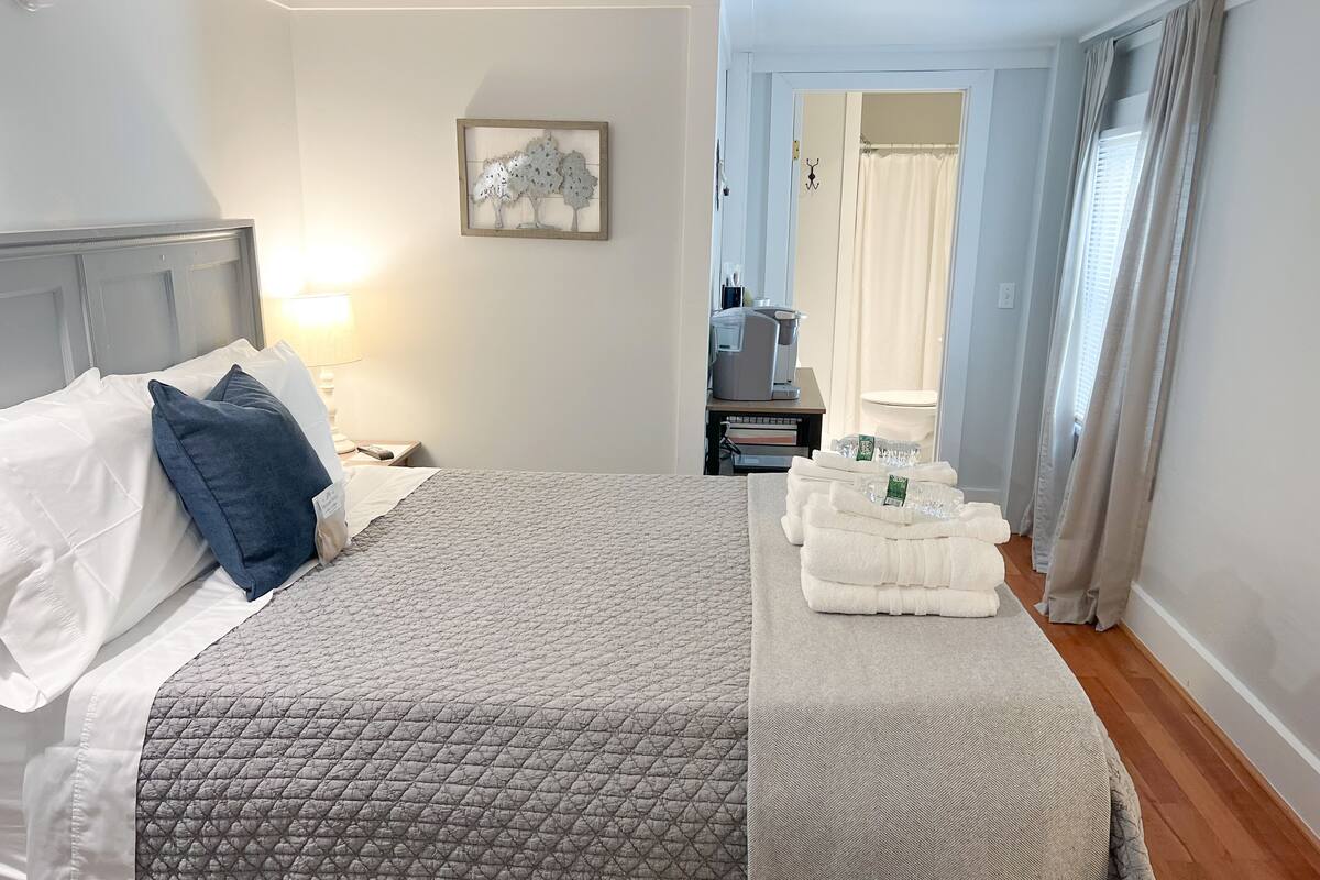 A neatly made bed with a quilted bedspread, a pillow, and a bedside table with a lamp. There is a framed artwork on the wall and a window with curtains.