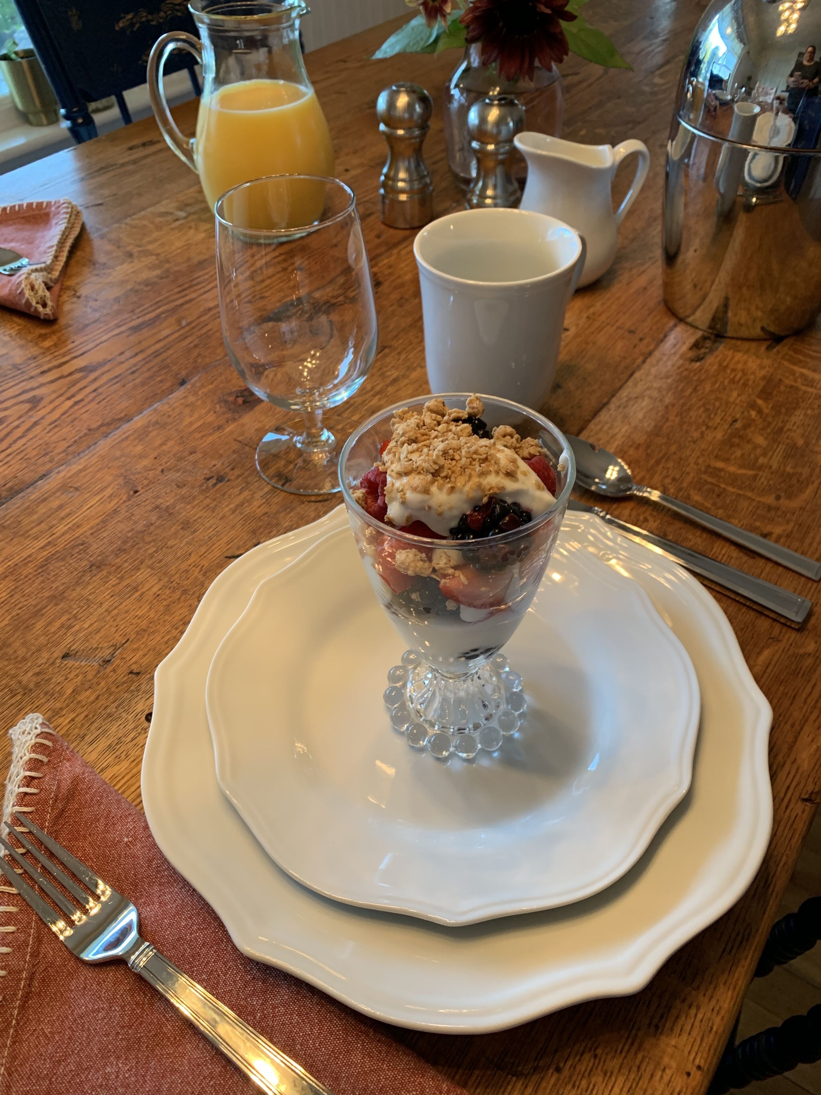 Morgenmadsbord med glasdessert på hvid tallerken, kande med appelsinjuice og kaffekande på et træbord.