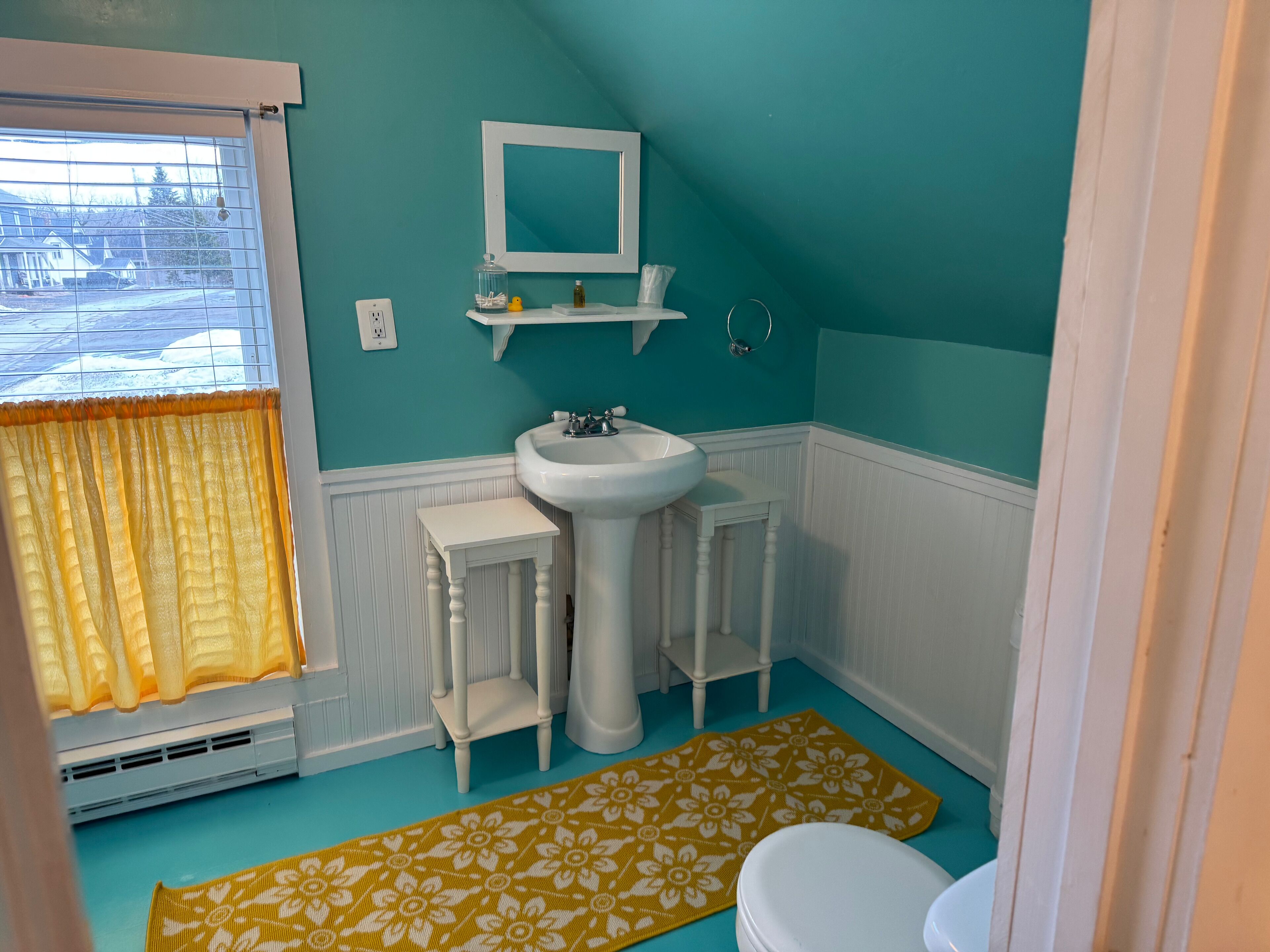 Un baño con un lavabo blanco, un espejo encima y un inodoro. Hay una alfombra estampada de color amarillo en el suelo.