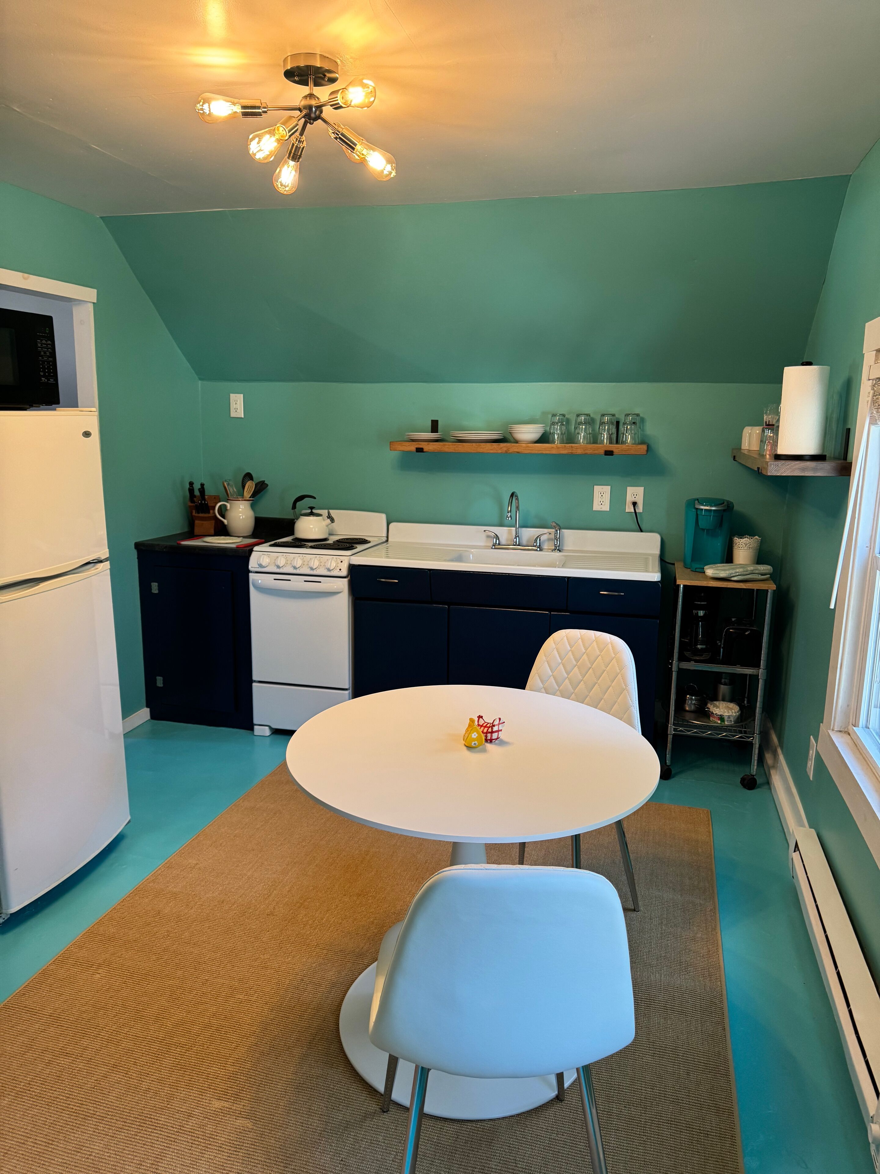 A compact kitchen with teal walls, a white refrigerator, a stove, and a sink. There is a small round table with a chair in the center.