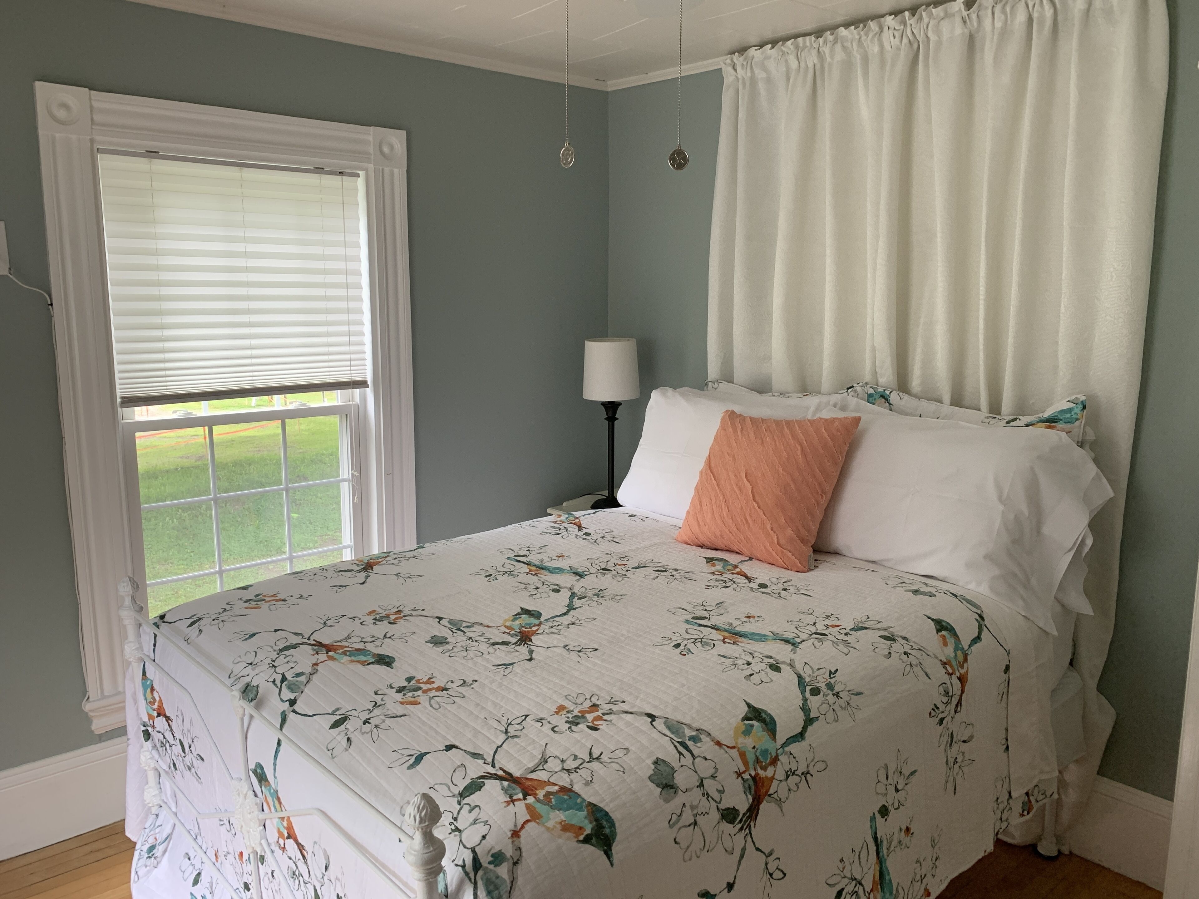 A bedroom with a bed, a window with blinds, a bedside lamp, and a view of the outdoors.