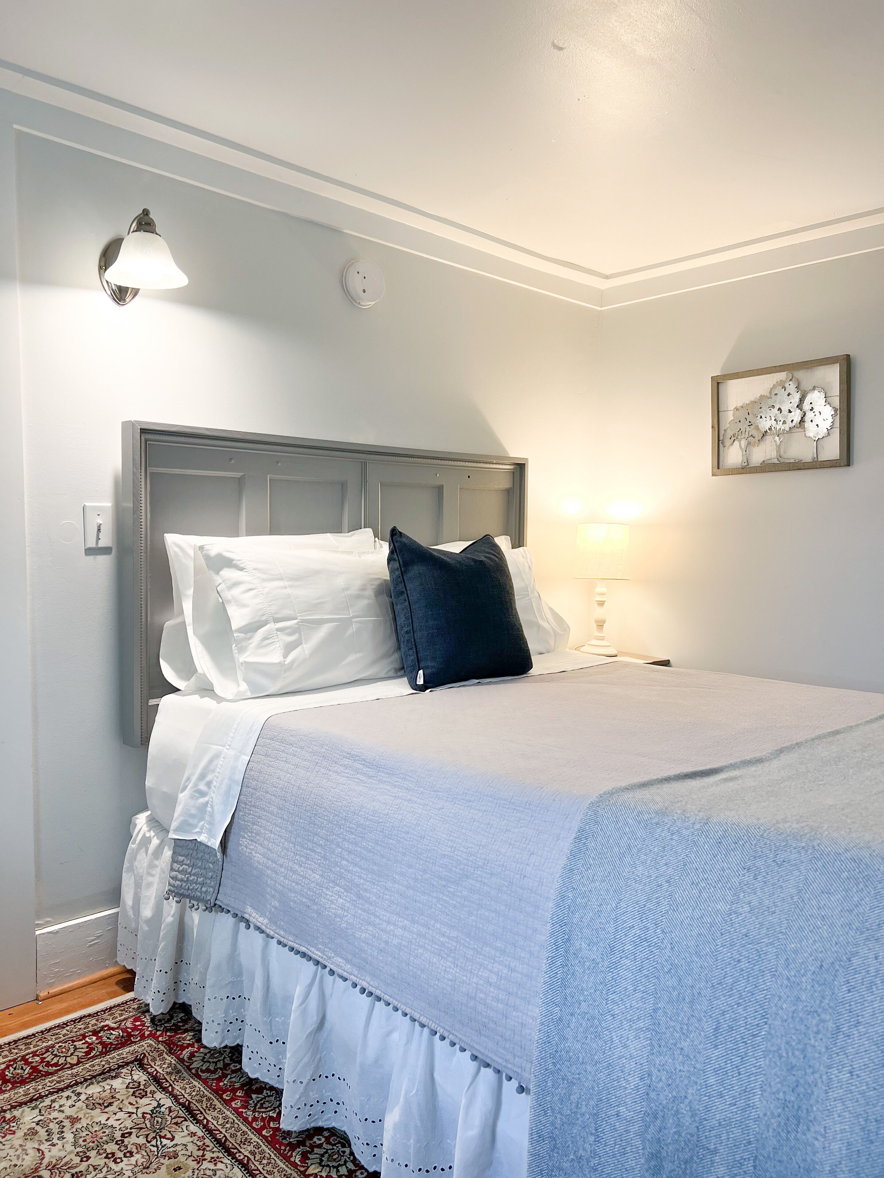 Un lit bien fait, avec une literie blanche et bleue, un oreiller bleu foncé, une lampe fixée au mur et une œuvre d’art encadrée accrochée au mur.