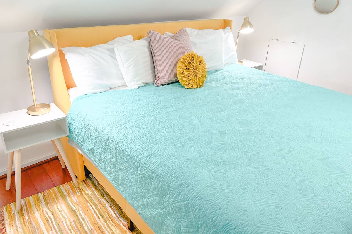 A neatly made bed with a light blue quilt, white pillows, and a decorative yellow flower. There is a bedside table with a lamp and a small shelf. The room has white walls and a wooden floor.