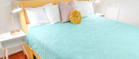 A neatly made bed with a light blue quilt, white pillows, and a decorative yellow flower. There is a bedside table with a lamp and a small shelf. The room has white walls and a wooden floor.