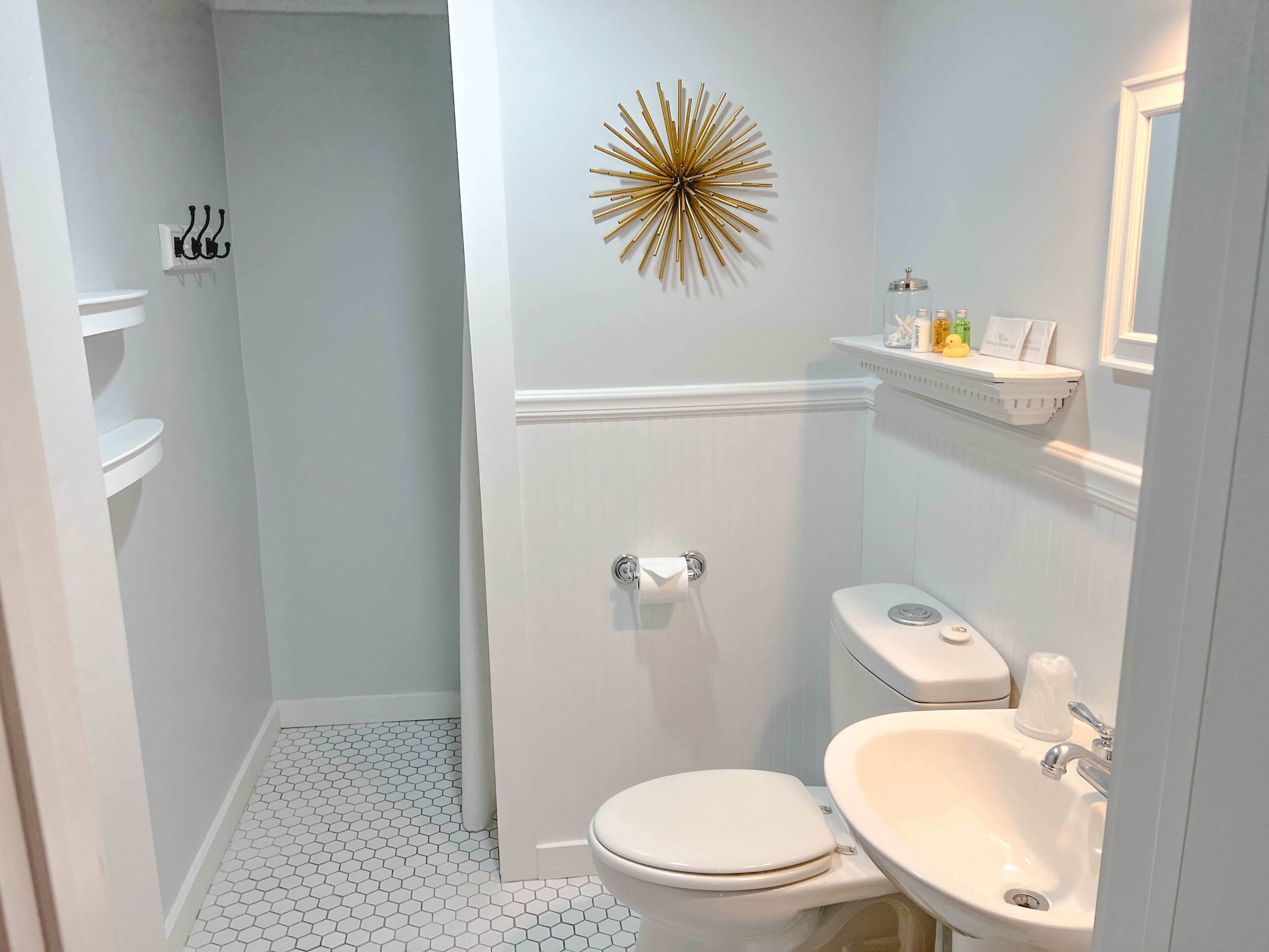 A bathroom with a toilet, sink, and shower. There is a decorative wall piece above the sink and a shelf with toiletries.