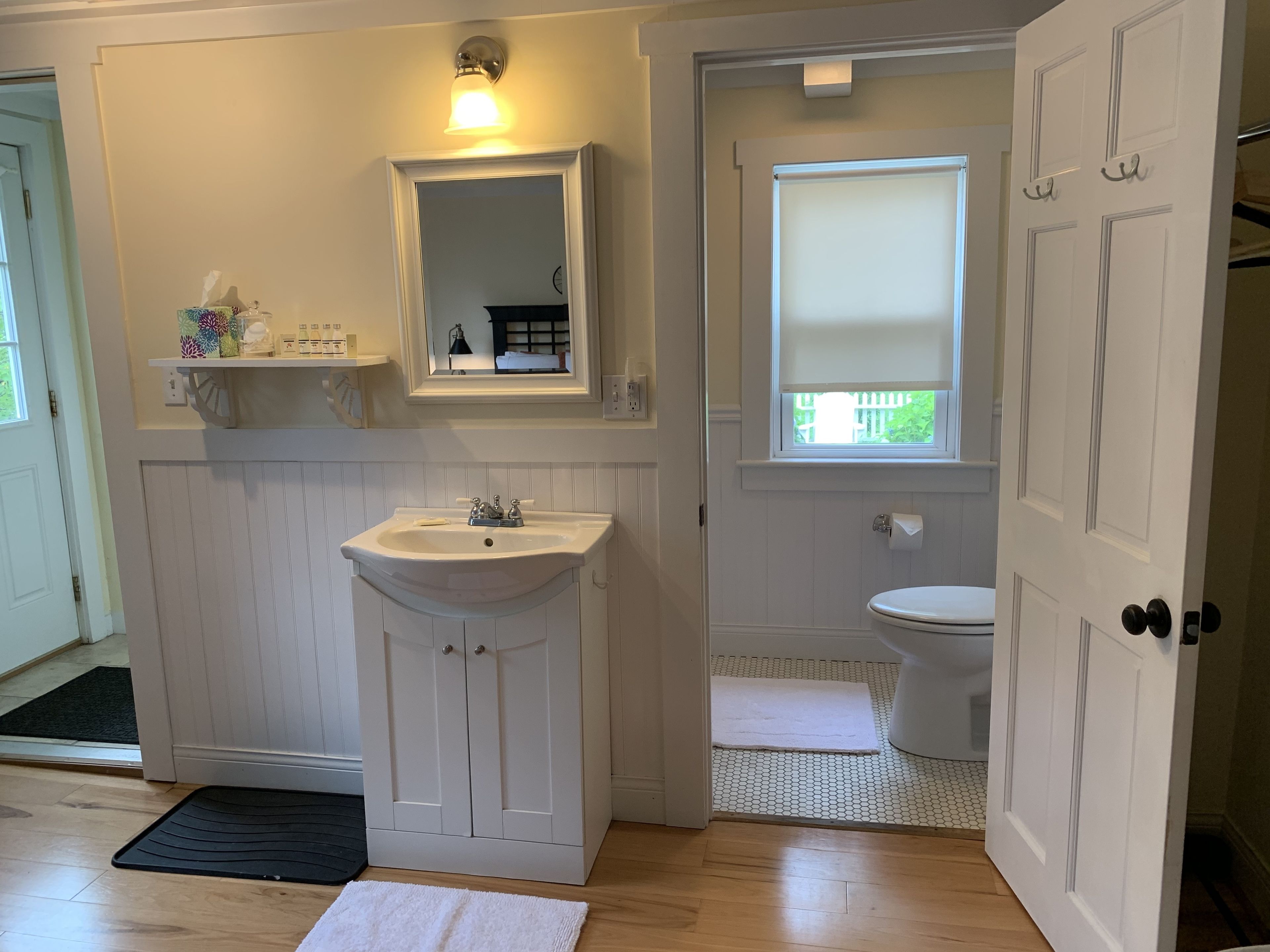 Un baño con lavabo blanco, espejo, inodoro, suelo de madera y una pequeña ventana con persiana.