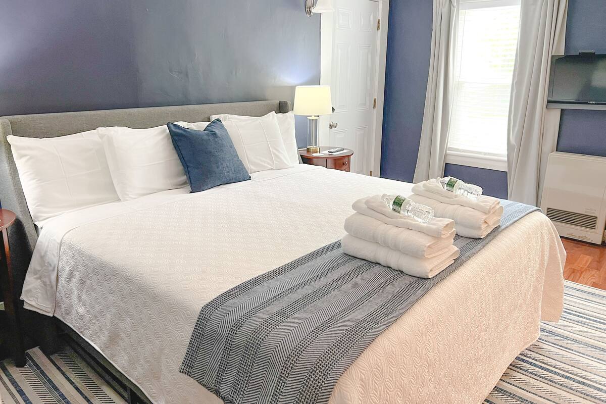 A neatly made bed with white and blue pillows, a striped blanket, and folded towels on a bed in a room with a large window and a TV on the wall.