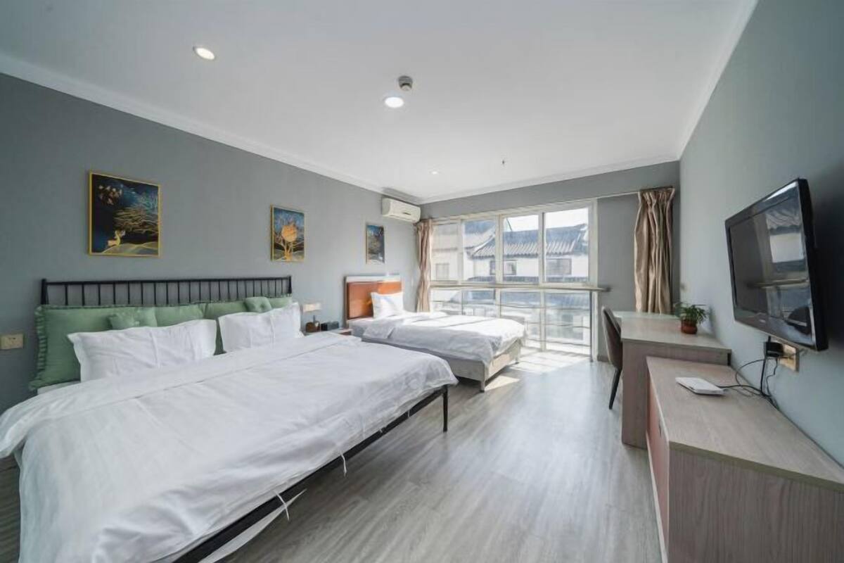 A modern hotel room with two beds, a desk, a TV, and large windows.