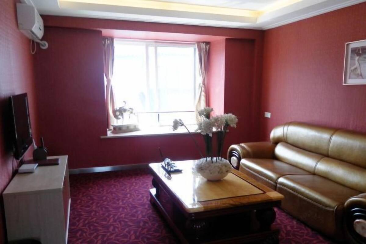 A room with a red wall, a brown leather sofa, a wooden coffee table with a vase of flowers, a flat-screen TV on a stand, and a window with curtains.