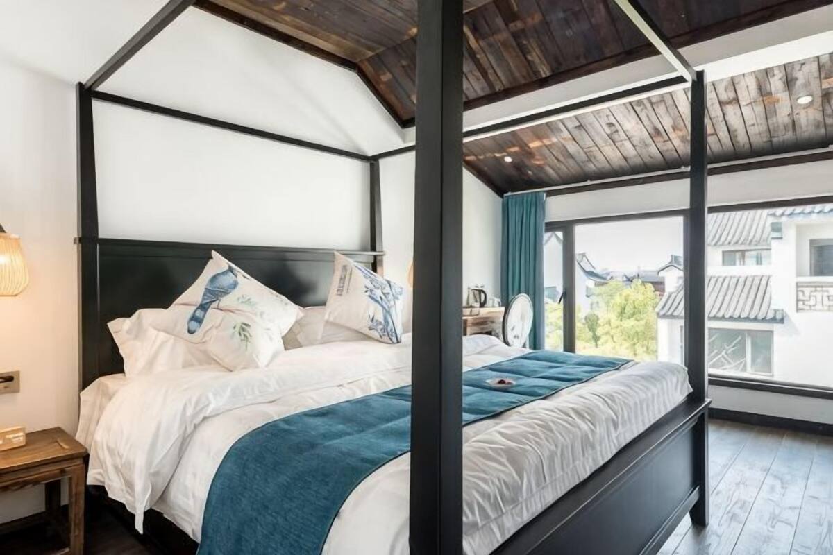 A four-poster bed with white bedding and a teal throw blanket, a wooden nightstand with a lamp, and a view of traditional buildings through large windows.