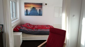 A bedroom with a single bed, a red chair, a painting of a pier, and a window with curtains. - High quality 1 1/2 room apt. with lake view and large terrace! (Allensbach)