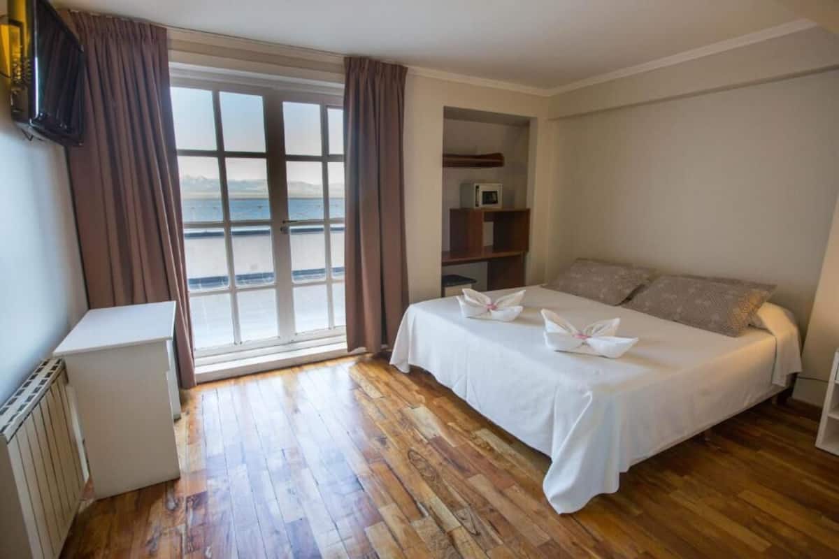 A bedroom with a wooden floor, a bed with white bedding, a small white desk, a shelf with a microwave, and a view of the sea through a large window.