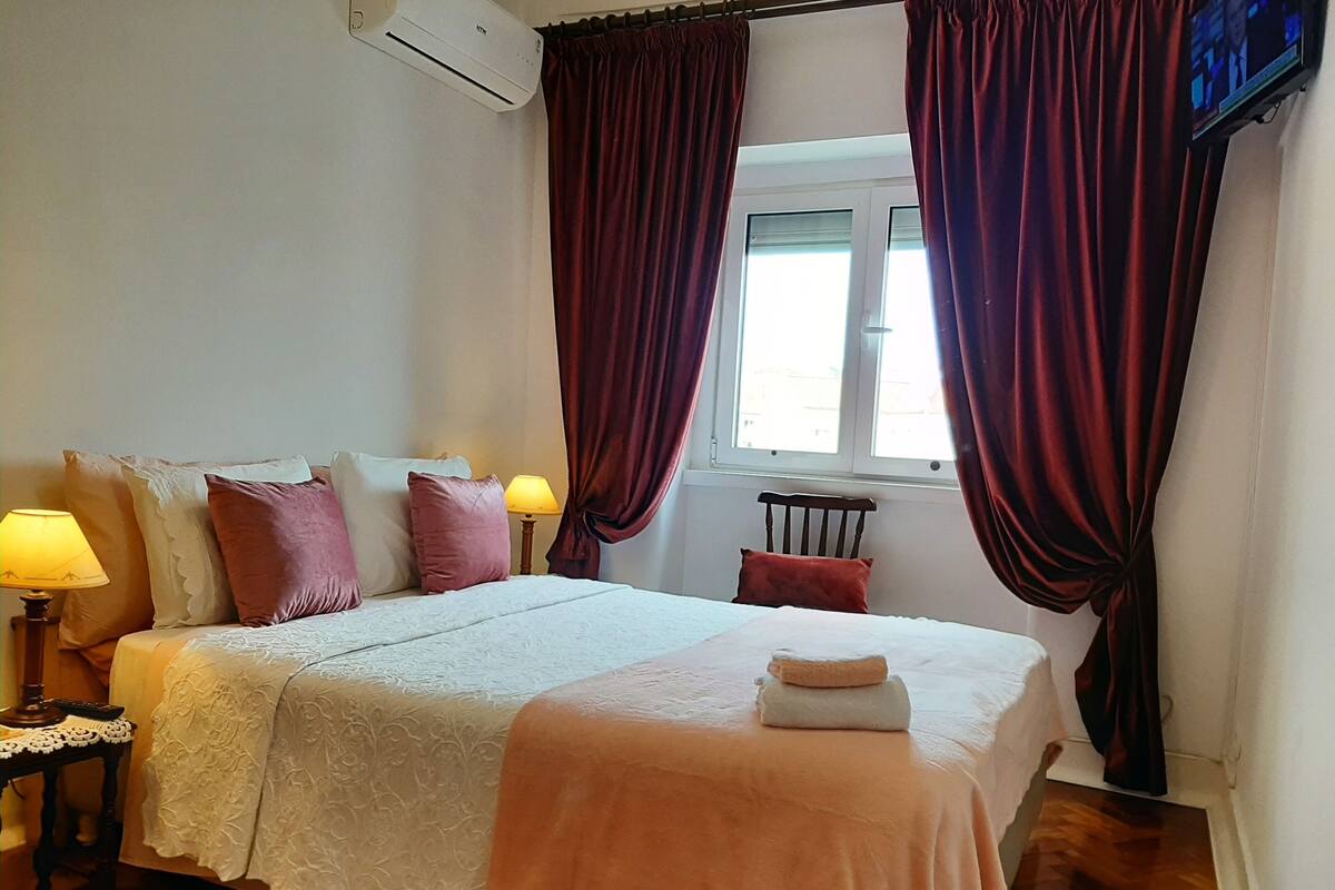 Een slaapkamer met een bed, kussens, een lamp, een televisie en een raam met gordijnen.