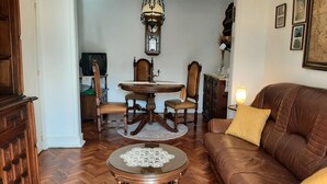Un salon chaleureux avec un canapé en cuir marron, une table basse ronde et une table à manger avec des chaises. Une horloge à coucou est accrochée au mur, et des tableaux encadrés sont disposés au-dessus du canapé.