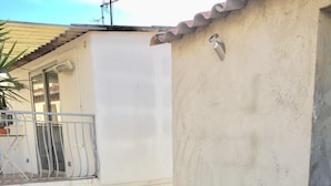 Deux fauteuils de jardin en bois, dotés de coussins blancs, sont placés sur une terrasse carrelée, juste à côté d’un bâtiment muni d’un balcon.