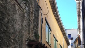 Une rue étroite bordée de bâtiments jaunes, de volets verts et d’un mur de pierre d’un côté.