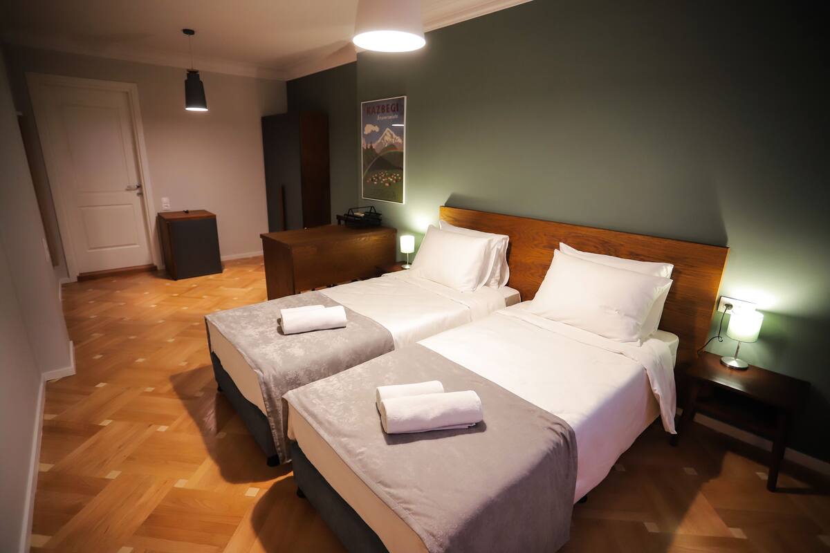 Habitación de hotel con dos camas, suelo de madera, un cartel en la pared y una mesita de noche con lámpara.