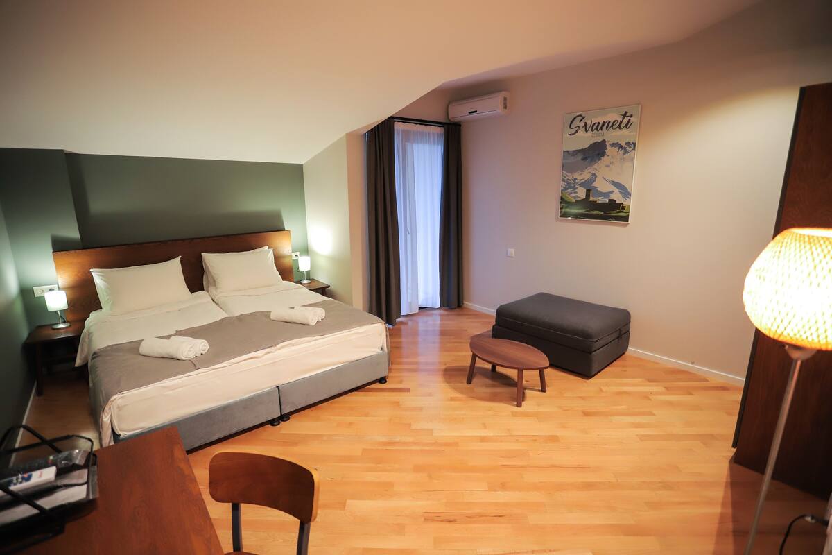 Habitación de hotel con cama, escritorio, silla, pequeño reposapiés, lámpara y un cartel en la pared.