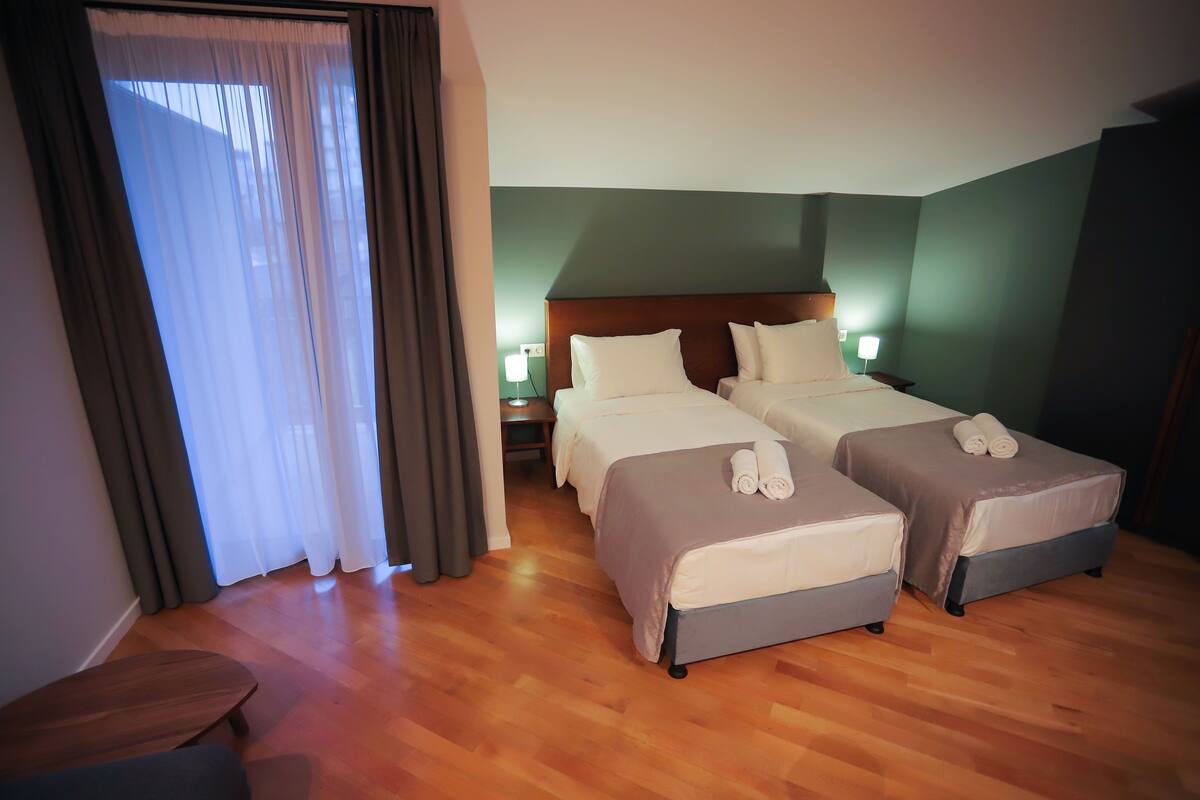 Habitación de hotel con dos camas, suelo de madera, mesita de noche con lámpara y ventana con cortinas.