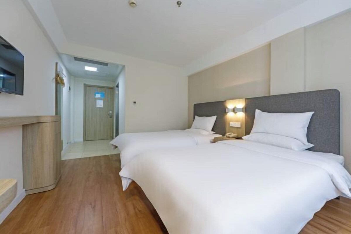 A hotel room with two beds, a wooden floor, a TV on the wall, and a door leading to another room.