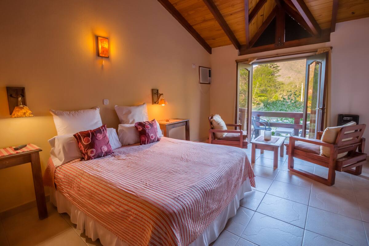 A bedroom with a large bed, a wooden ceiling, a balcony with chairs, and a view of greenery.