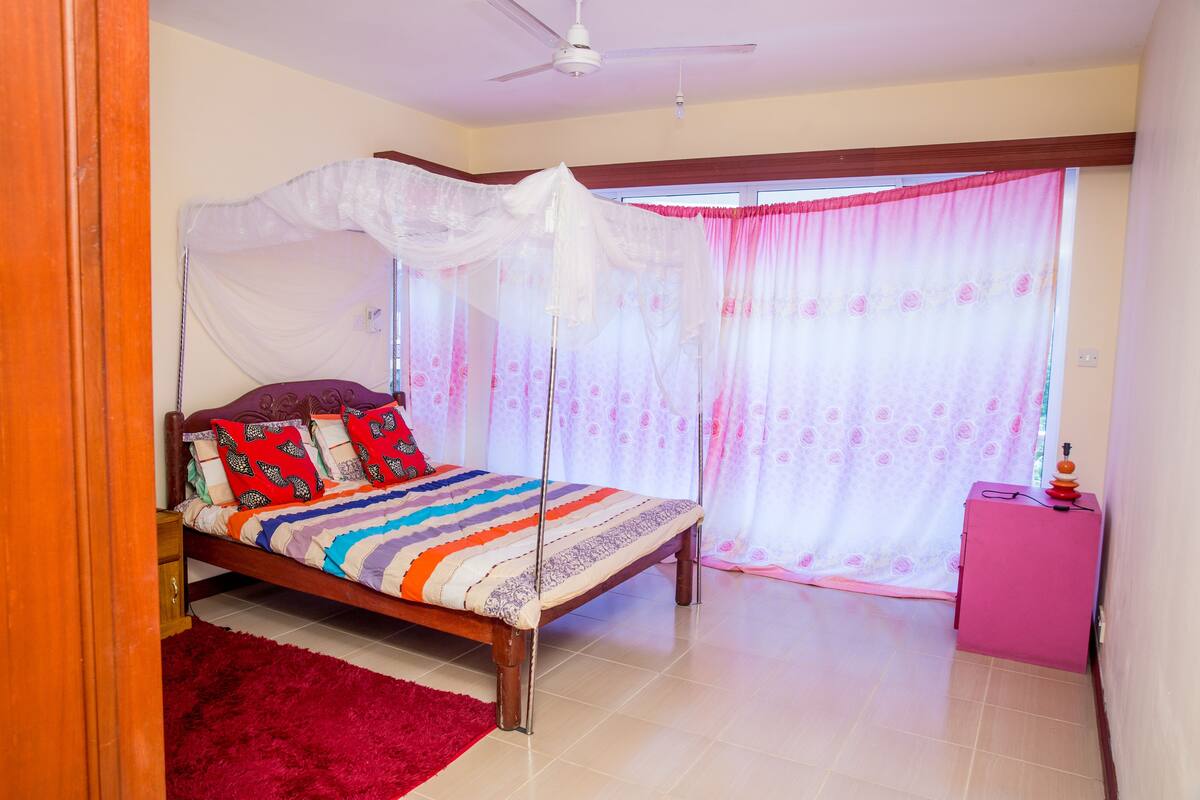 A bedroom with a bed, a ceiling fan, a window with curtains, and a small pink cabinet.
