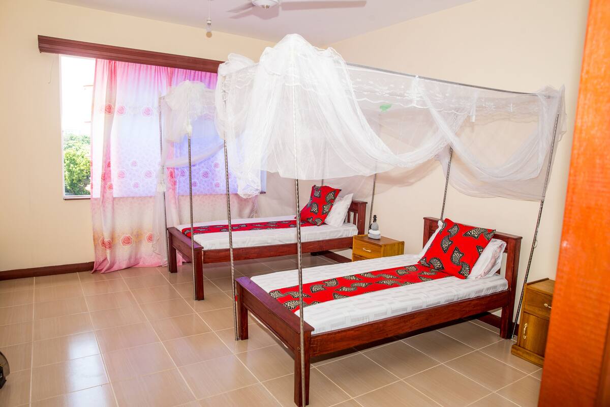 A room with two wooden beds, each with a canopy. There is a window with pink curtains, a wooden nightstand, and a small table with a lamp.
