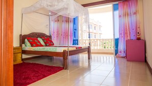 A bedroom with a bed, a red rug, a balcony with curtains, and a view of buildings. - Safiri Kenya Home (Mombasa)